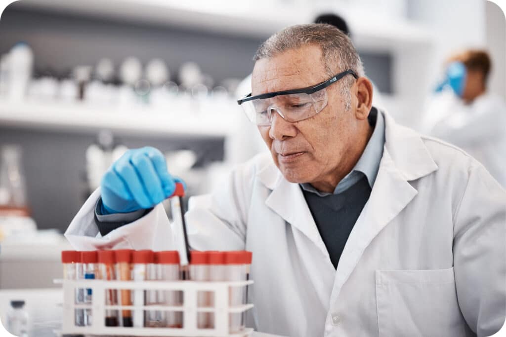 A medical professional with blue gloves and protection glasses pulls a test tube out of a test tube tray.
