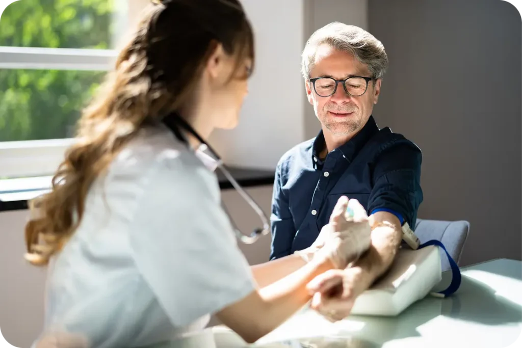 A healthcare professional prepares to draw blood from a seated man’s arm with sunlight coming through a window.